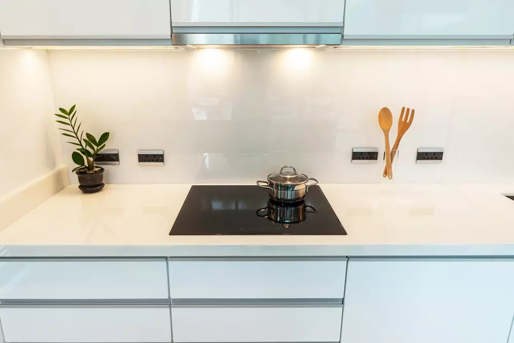 Cozinha moderna com bancada branca, cooktop de indução, panelas, utensílios de madeira e uma planta de interior mostrando um ambiente limpo e organizado.