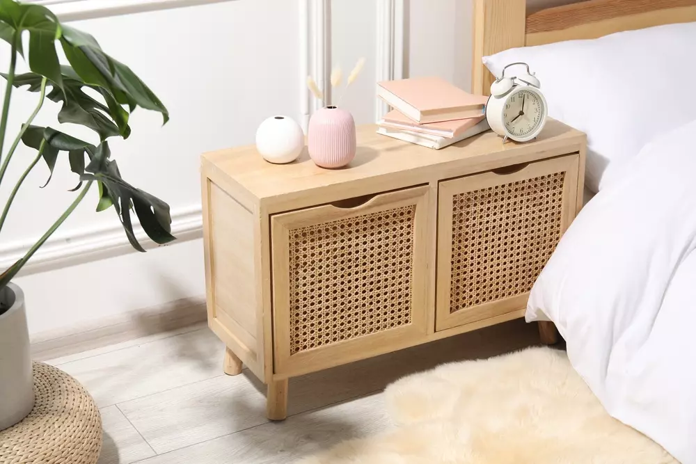 Cômoda de madeira rústica com gavetas de rattan ao lado da cama em um quarto decorado com estilo minimalista, incluindo plantas, despertador, livros e objetos decorativos.