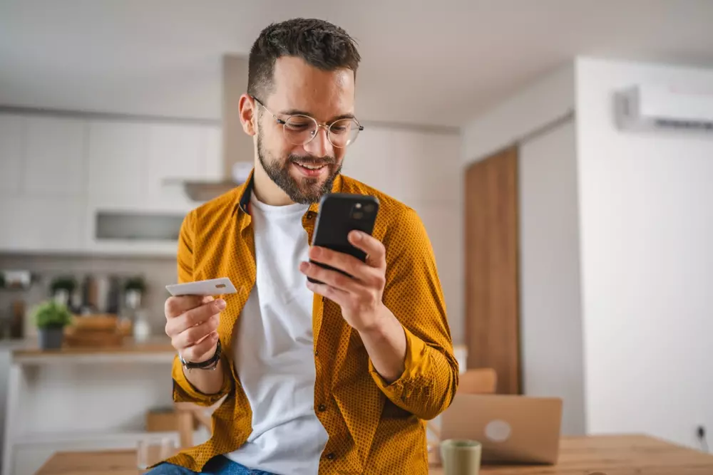 Homem sorridente utilizando smartphone e cartão digital no ambiente de casa, representando estratégias de juntar dinheiro usando plataformas digitais e economia doméstica