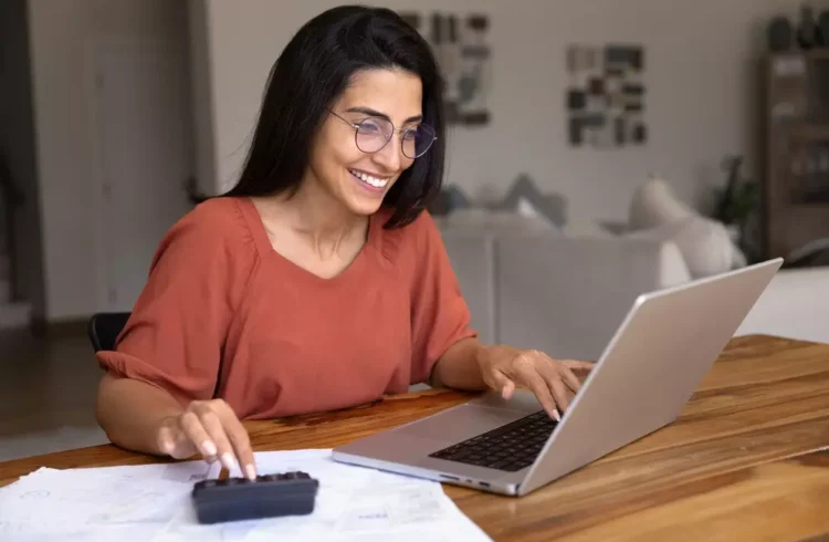 Mulher sorridente usando laptop e calculadora em uma mesa, promovendo a ideia de pagar-se primeiro para melhor saúde financeira.