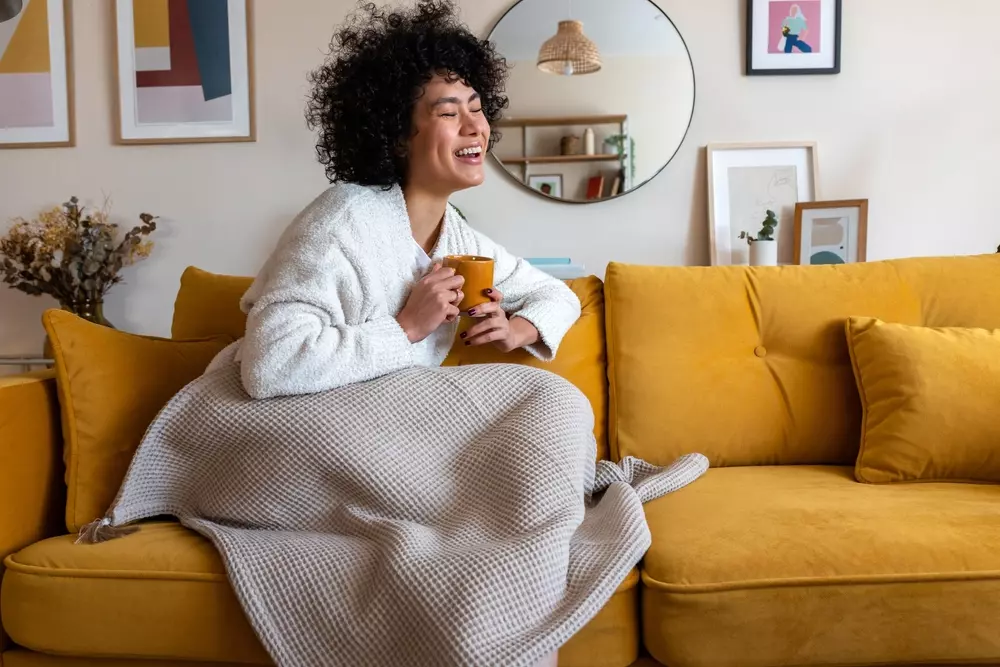 Mulher sorridente sentada em sofá amarelo, confortável com uma xícara, transmitindo sensação de aconchego e bem-estar em casa.