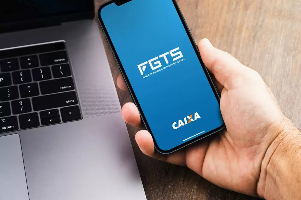 Pessoa segurando smartphone mostrando o aplicativo do FGTS e Caixa, soluções de pagamento digital, próximo ao laptop em uma mesa de madeira.