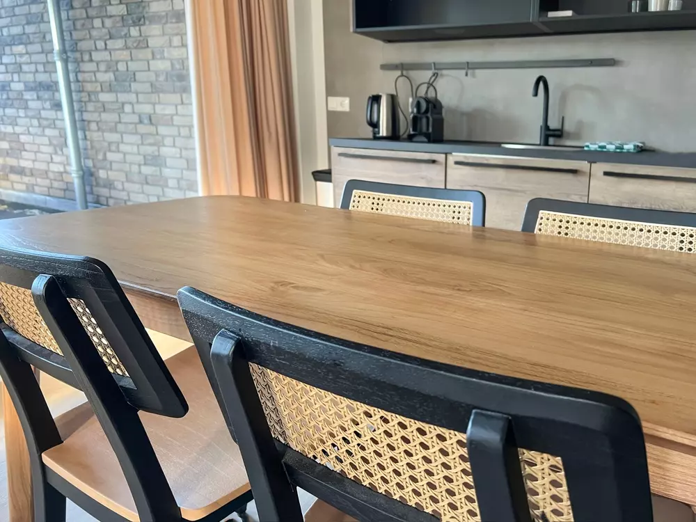 Uma sala de jantar moderna com mesa de madeira oval, cadeiras com assento de vime e uma cozinha de fundo com bancada, torneira e eletrodomésticos, decorada em estilo contemporâneo.