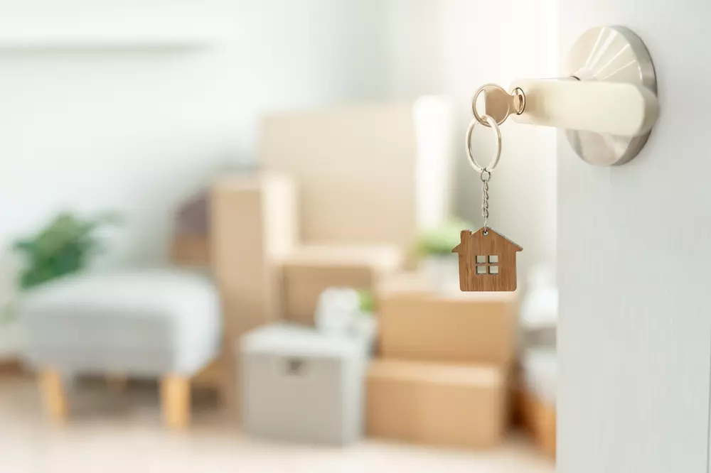 Chave de porta decorada com uma miniatura de casa pendurada na maçaneta, simbolizando mudança ou novo lar, em um ambiente de sala de estar com caixas de mudança.