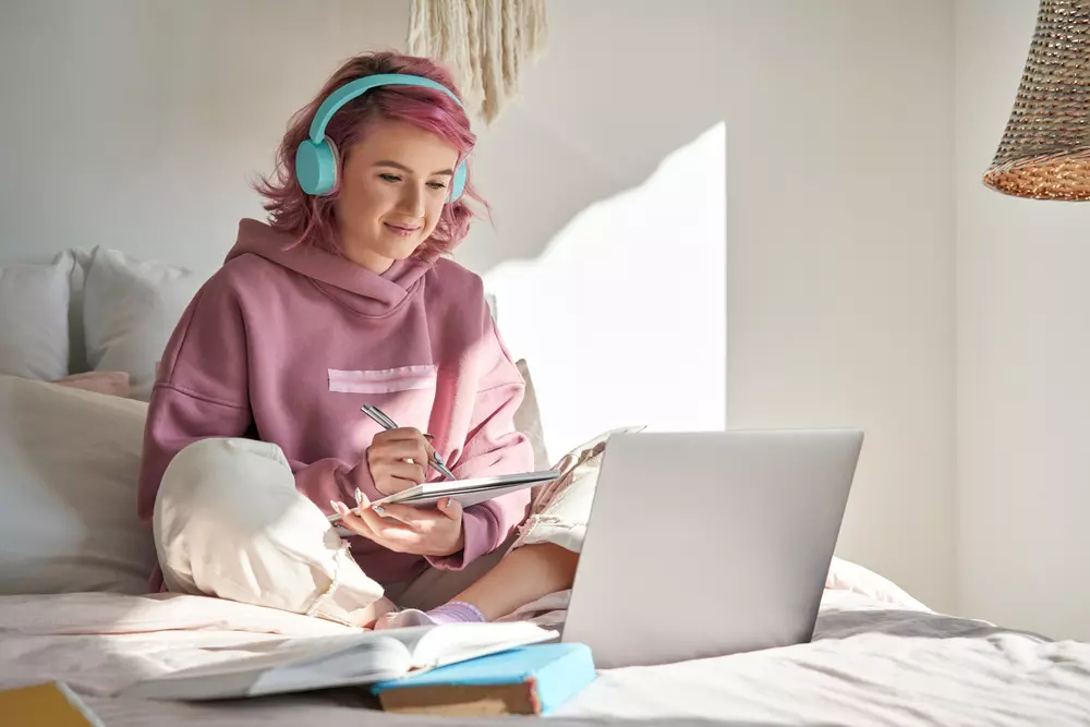Jovem de cabelo rosa sentada em sua cama estudando com headphones, caderno e laptop. Representando a geração do quarto.