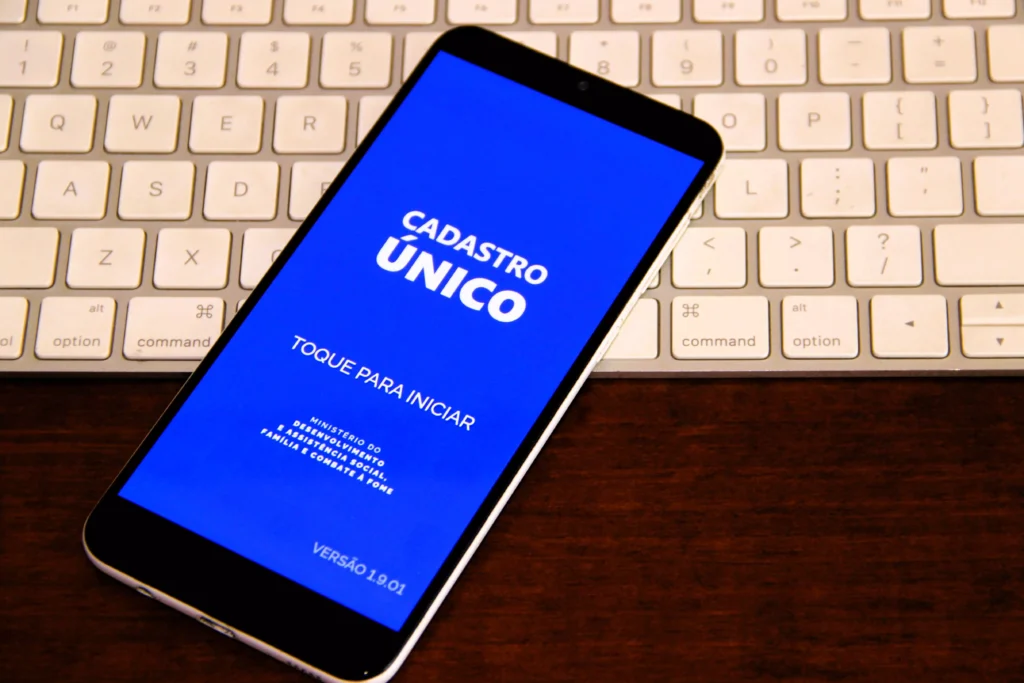 Um aparelho celular posicionado em cima de um teclado de computador branco. Na tela do celular o sistema de Cadastro Único do governo. Estar inscrito e com os dados familiares atualizados no CadÚnico é um dos requisitos para o programa Minha Casa Minha Vida