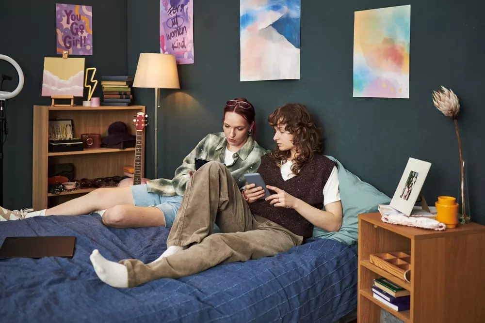 Duas jovens sentadas na cama de um quarto decorado com quadros abstratos, uma usando óculos e ambas usando roupas casuais, enquanto olham para seus celulares. Representando a geração do quarto.