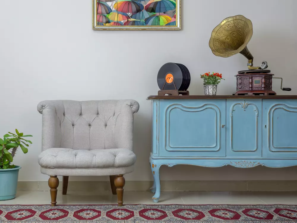 Sala com cadeira confortável, rádio antigo, vitrola, vaso de plantas, quadro de guarda-chuvas coloridos e móvel azul vintage, no estilo do meme à moda.