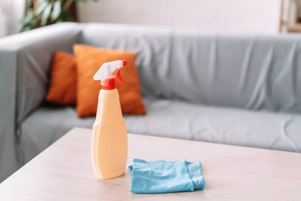 Produto de limpeza com frasco spray e pano em mesa de centro na sala, fundo com sofá cinza e almofadas laranja, ideal para manter a limpeza doméstica.