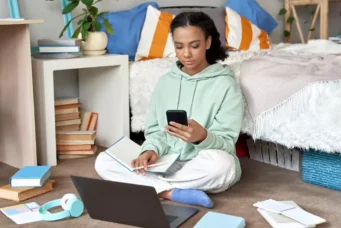 Jovem estudando em seu quarto, sentada no chão com livros, laptop e smartphone em ambiente de quarto organizado com cores vibrantes. Representando a geração do quarto