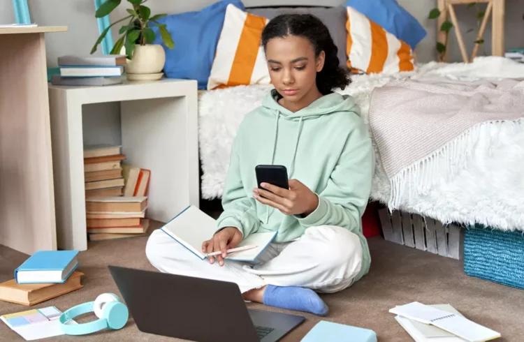 Jovem estudando em seu quarto, sentada no chão com livros, laptop e smartphone em ambiente de quarto organizado com cores vibrantes. Representando a geração do quarto