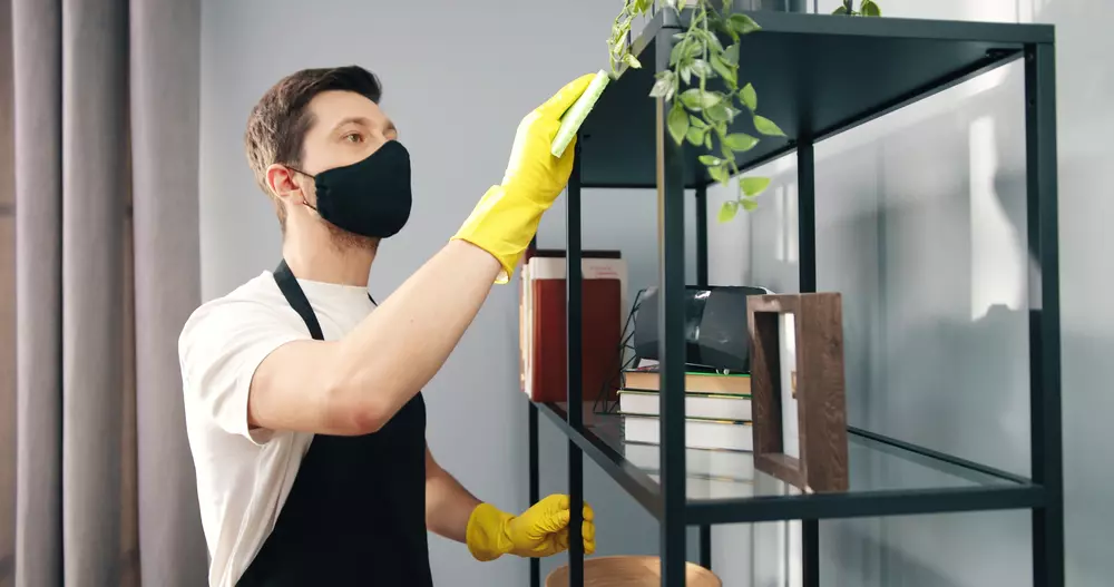 Homem organizando e limpando uma estante com livros e plantas, usando máscara de proteção e luvas, em ambiente interno.