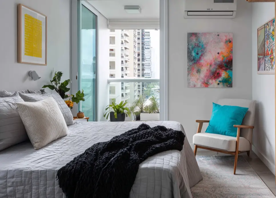 Quarto com varanda integrada, decoração moderna com quadros coloridos, plantas e vista de prédios altos ao fundo
