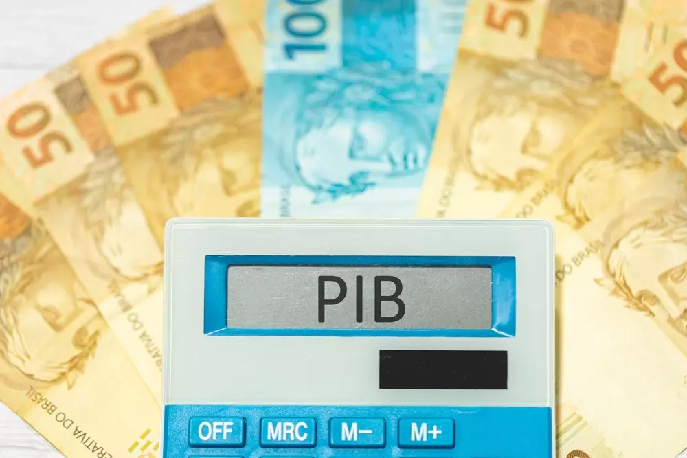 Notas de 50 e 100 Reais dispostas em cima de uma mesa, com uma calculadora posicionada acima delas escrito "PIB" no visor. Representando as cidades mais ricas do Mato Grosso do Sul
