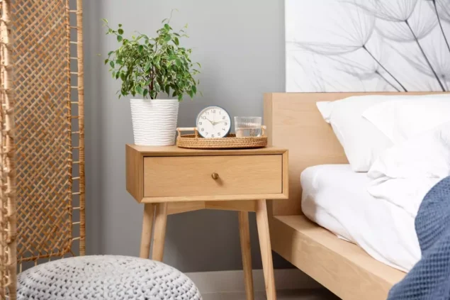 Uma mesa de cabeceira de madeira ao lado de uma cama com roupa branca, decorada com um vaso de plantas, um relógio, um copo de água e uma bandeja. Ambiente aconchegante e moderno.