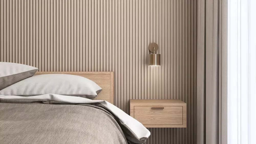 Uma mesa de cabeceira suspensa de madeira ao lado de uma cama com roupa de cama cinza e branco, decorada com parede de painel ripado e cortinas brancas ao fundo.