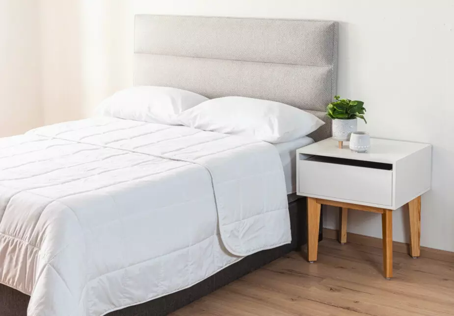 Quarto de casal com cama confortável, roupa de cama branca, cabeceira estofada cinza com pés de madeira clara, minimalista com planta, ideal para ambientes modernos e confortáveis.