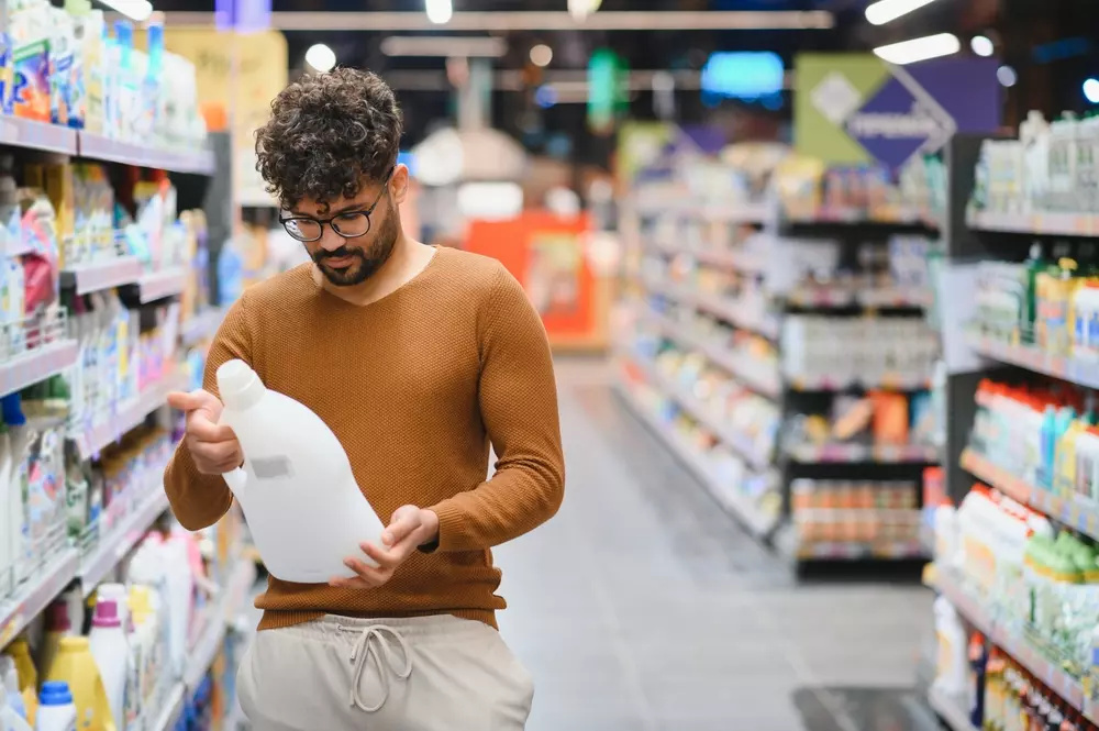Homem no corredor de produtos de limpeza de um supermercado lendo o rótulo de um produto. Um dos erros de limpeza é não ler o rótulo antes do uso.