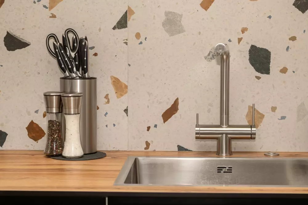 Cozinha com uma bancada de madeira clara, topo de pia de aço inox, utensílios de cozinha em um cilindro de metal e um conjunto de três potes de tempero, tudo com bagunça intencional na parede decorativa de fundo.