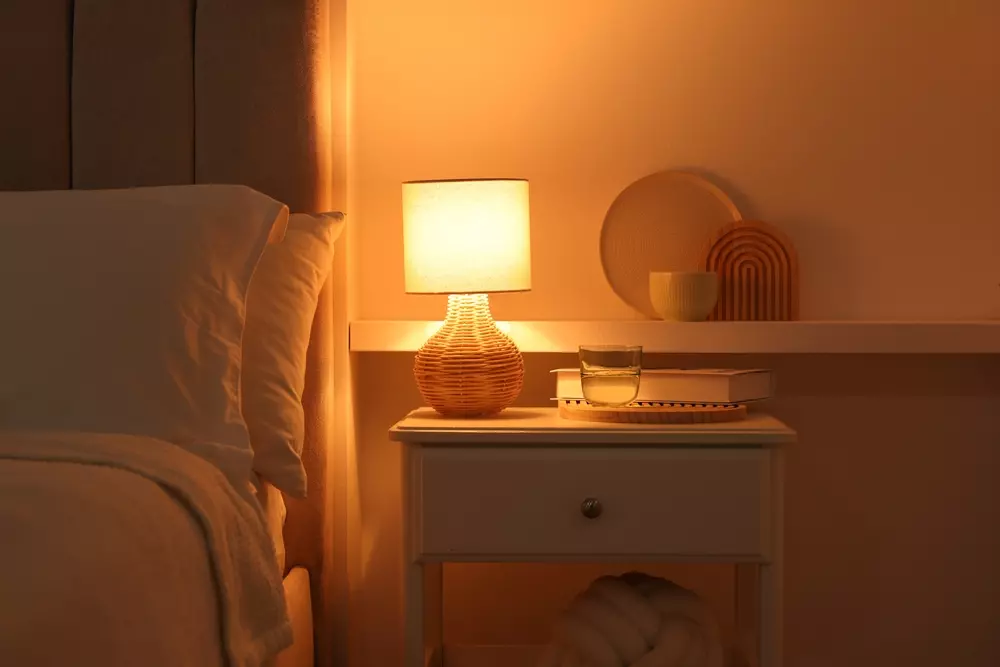 Detalhe de uma mesa de cabeceira com profundidade, iluminada por uma luminária aconchegante, com objetos decorativos e uma cama ao lado.