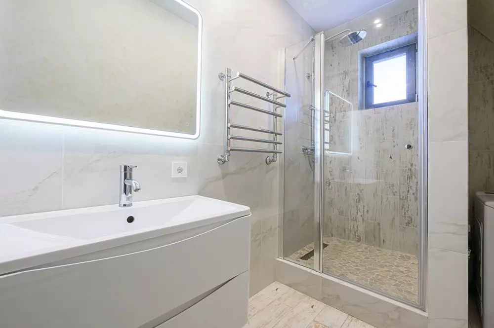 Banheiro moderno com toalheiro elétrico próximo ao box de vidro e espelho iluminado, ideal para uso doméstico e fácil de instalar.