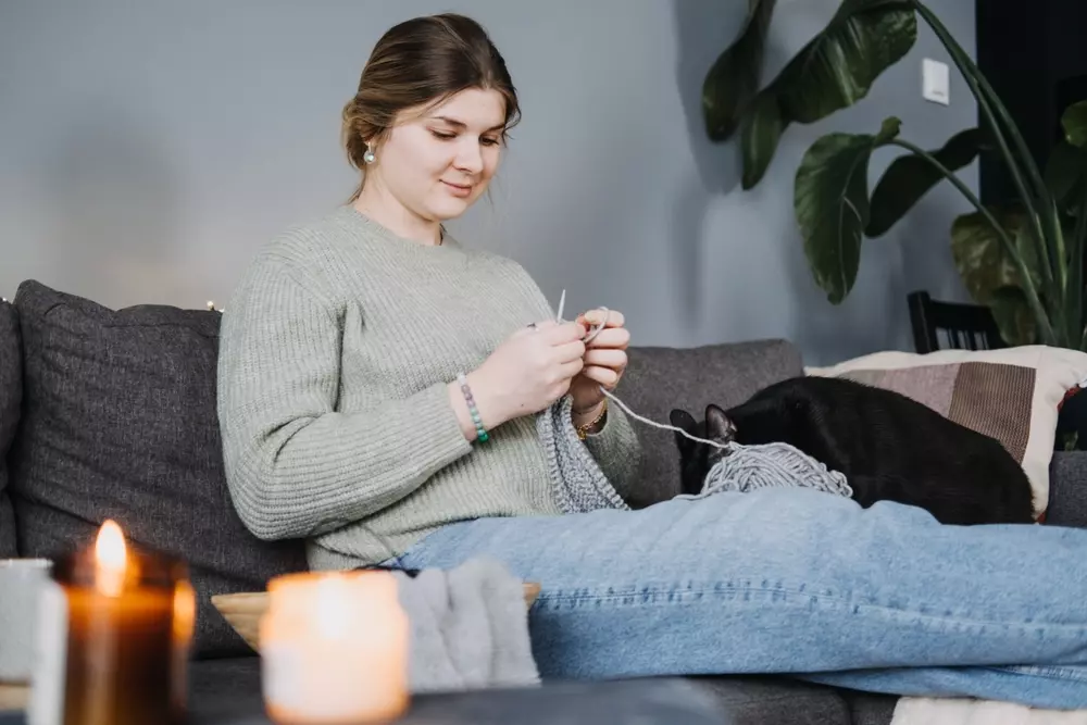 Mulher relaxando no sofá com uma postura de slow living e uma pequena bagunça boa ao seu redor, incluindo uma bola de lã e uma planta de interior, refletindo o essencialismo do ritmo de vida lento.