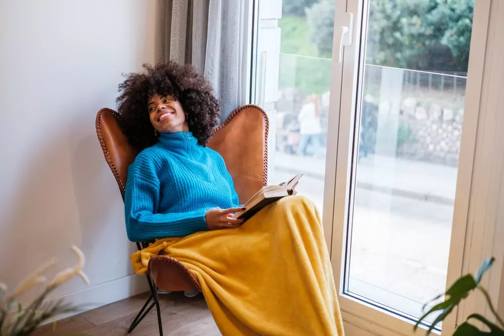 Mulher sorridente lendo um livro, sentada confortavelmente junto à janela, simbolizando sustentabilidade e valorização do bem-estar pessoal.