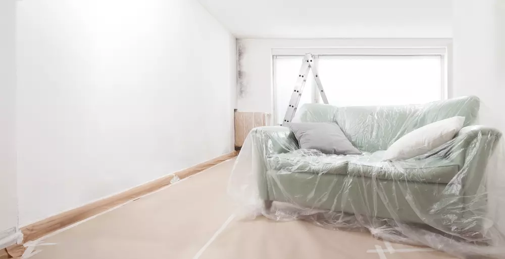 Sala de estar moderna em reforma, com sofá coberto em plástico, escada de madeira ao fundo, acabamento em tons claros e janela grande permitindo entrada de luz natural.