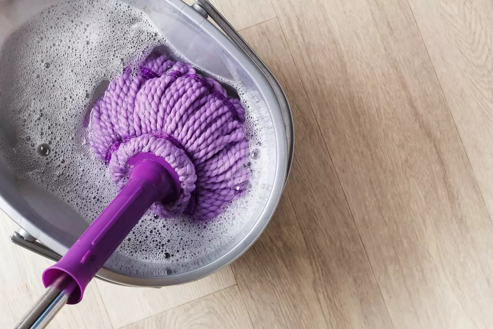 Balde com esfregão lavando piso de madeira com água e espuma, ferramenta de limpeza doméstica eficiente.