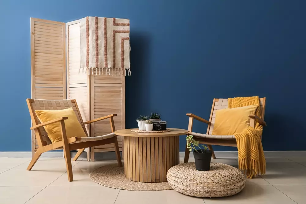 Biombo decorativo com textura adicionada, ambiente com cadeiras de madeira, almofadas amarelas, mesa de centro rústica, e elementos decorativos em espaço interno com parede azul.