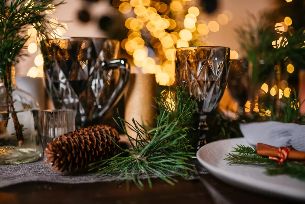 Centro de mesa de Natal com ramos verdes, pinhas e taças decorativas em uma mesa festiva, com luzes de Natal ao fundo criando atmosfera aconchegante
