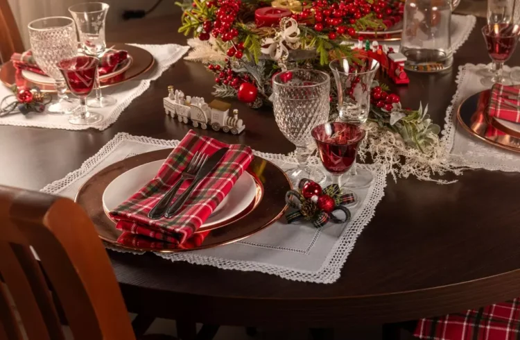 Decoração de centro de mesa de Natal com arranjo de folhas verdes, galhos de azevinho, bolas vermelhas, caixas decorativas vermelhas, taças de vidro com vinho, pratos, talheres e guardanapos com padrão xadrez vermelho e verde.