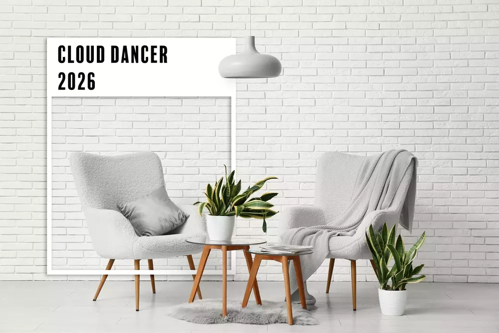 Sala com duas poltronas brancas, mesa de centro de madeira, plantas em vasos brancos e quadro com a frase 'Cloud Dancer 2026' fazendo referência à cor escolhida pela Pantone.