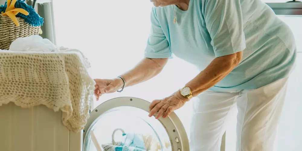 Mulher idosa carregando a porta da máquina de lavar roupas na cozinha, promovendo redução de esforço na lavanderia.