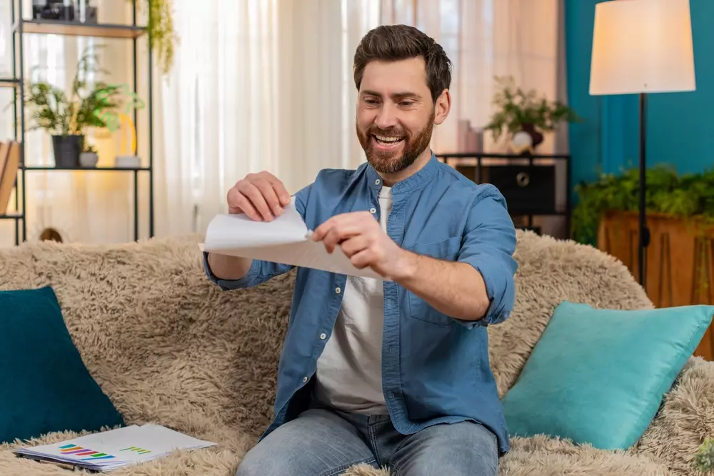 Jovem no sofá de casa, sorrindo e rasgando um boleto desnecessário em pedaços com alívio.