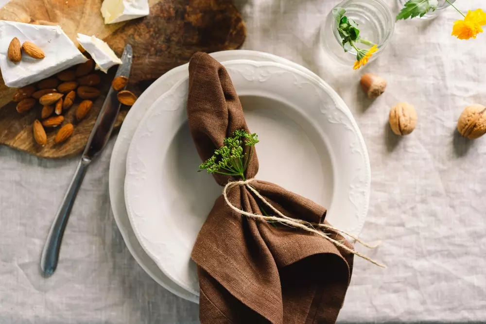 Guardanapo de linho marrom com amarração delicada e toques verdes, sobre um prato branco em mesa de jantar elegante, decorado com flores e nozes.