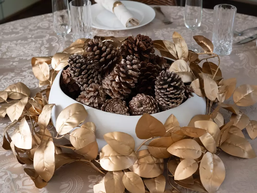Conjunto de pinheiros decorativos dourados e uma tigela branca cheia de pinhas naturais na mesa de jantar, criando uma decoração elegante e natalina.