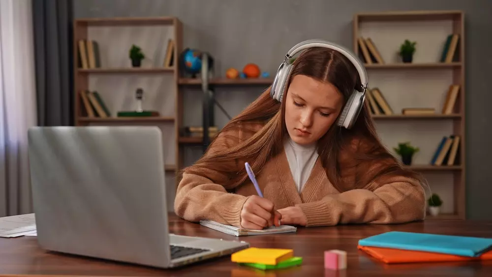Menina adolescente estudando com fone de ouvido, ao lado de um laptop e materiais de papelaria, ideal para ouvir músicas para foco e concentração em casa ou escritório.