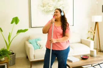 Pessoa feliz ouvindo músicas para foco usando fones de ouvido em casa enquanto limpa a casa, promovendo bem-estar e concentração.