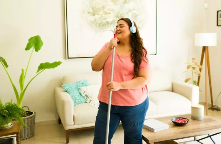 Pessoa feliz ouvindo músicas para foco usando fones de ouvido em casa enquanto limpa a casa, promovendo bem-estar e concentração.