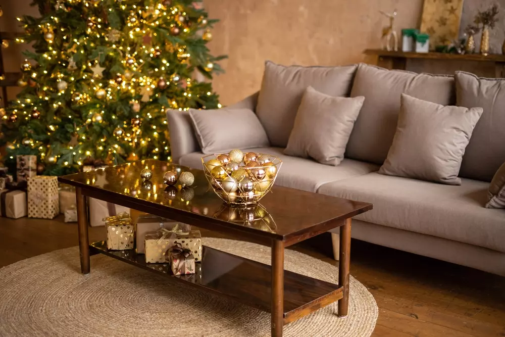 Sala de Natal decorada com árvore iluminada, sofá cinza com almofadas, mesa de centro de madeira com bolas douradas, presentes de Natal e decoração aconchegante.