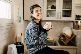 Mulher sorridente em uma Tiny House, segurando uma xícara de café na cozinha compacta e sustentável, demonstrando o estilo de vida minimalista e ecológico.