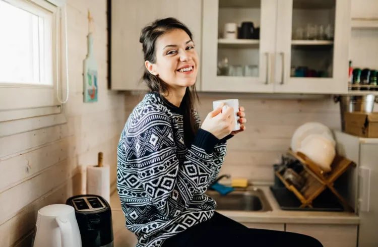 Mulher sorridente em uma Tiny House, segurando uma xícara de café na cozinha compacta e sustentável, demonstrando o estilo de vida minimalista e ecológico.