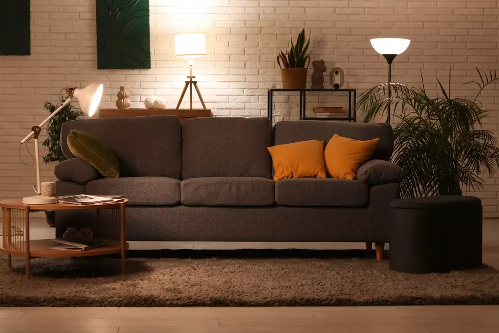 Sala moderna decorada com sofá cinza, almofadas amarelas, iluminação ambiente, plantas decorativas e uma parede de tijolos brancos que valorizam o espaço e criam uma atmosfera aconchegante.