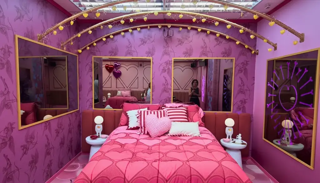 Quarto BBB26 rosa decorado com detalhes dourados, paredes roxas, espelhos, iluminação neon, cama com almofadas rosa e objetos decorativos