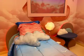 Quarto com decoração de nuvens, incluindo cama com roupa de cama azul, almofadas em formato de nuvem, painel com céu e nuvens, ambiente aconchegante para sonhos de voar no BBB26.