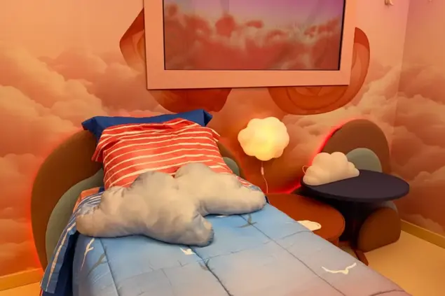 Quarto com decoração de nuvens, incluindo cama com roupa de cama azul, almofadas em formato de nuvem, painel com céu e nuvens, ambiente aconchegante para sonhos de voar no BBB26.