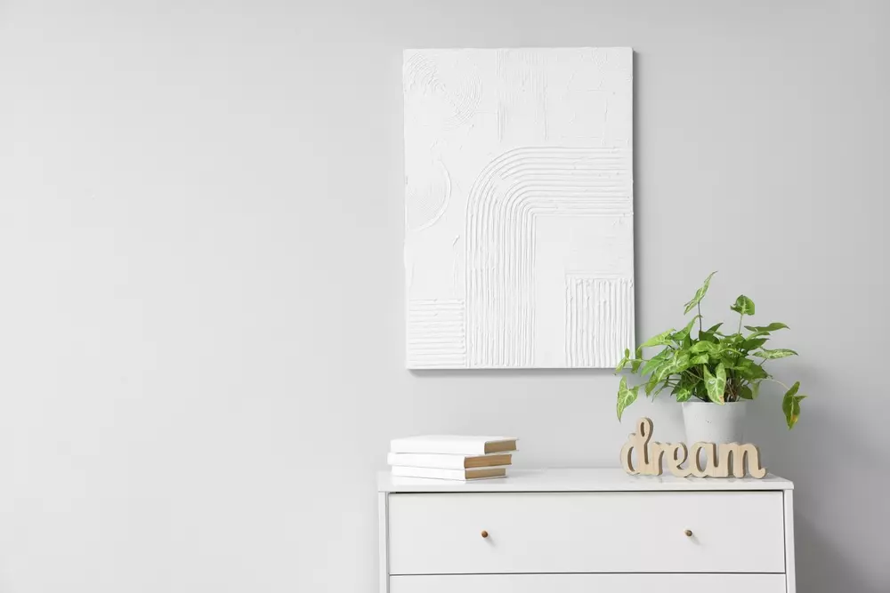 Decoração de ambiente com sofisticação em off-white, destaque para quadro branco com textura e uma planta em vaso sobre móvel branco.