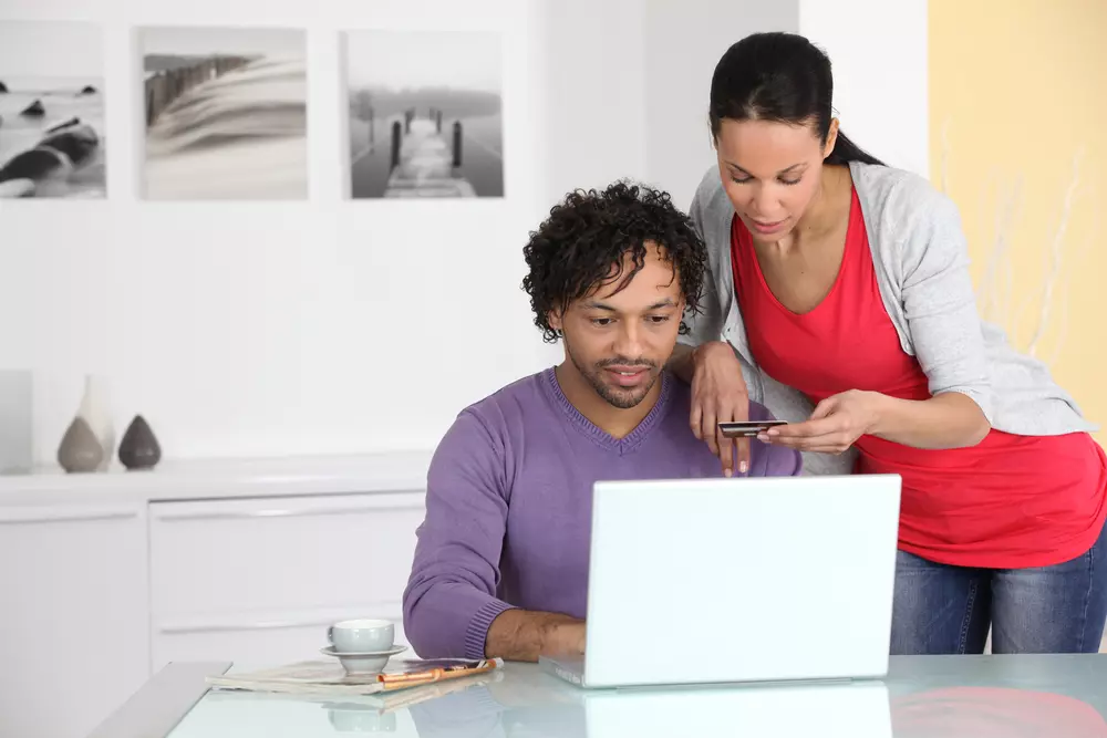 Homem e mulher usando laptop e smartphone juntos em ambiente doméstico moderno, com decoração minimalista e fotos na parede ao fundo.