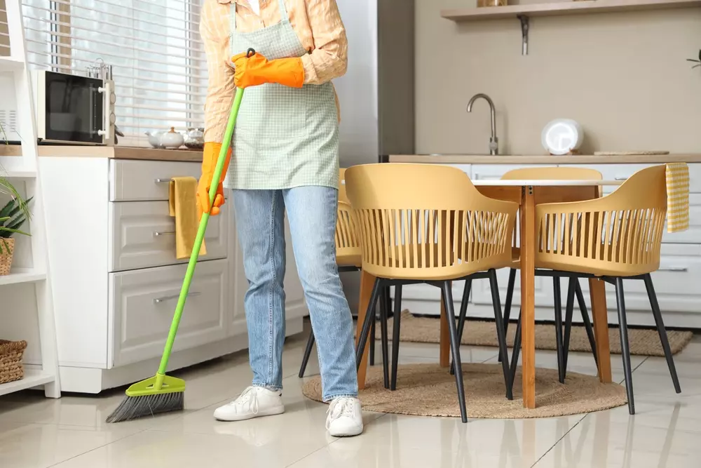 Pessoa fazendo limpeza na cozinha com vassoura, avental e luvas de borracha, ambiente moderno com mesa de jantar de madeira e cadeiras.